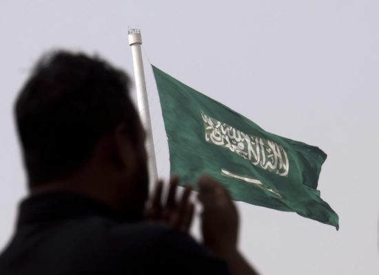 A man prays at an open air makeshift mosque in front of a giant Saudi Flag in Jiddah, Saudi Arabia, June 21, 2017. Saudi Arabia is inching towards changes that govern its national anthem and green flag, which is emblazoned with a sword and inscribed with Islam’s creed. State-owned media reported the kingdom’s un-elected consultative Shura Council voted in favor of changes late Monday, Jan. 31, 2022